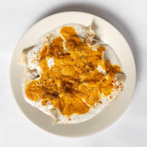Overhead view of manti dumplings covered in white yogurt sauce and orange chili oil, garnished with herbs.
