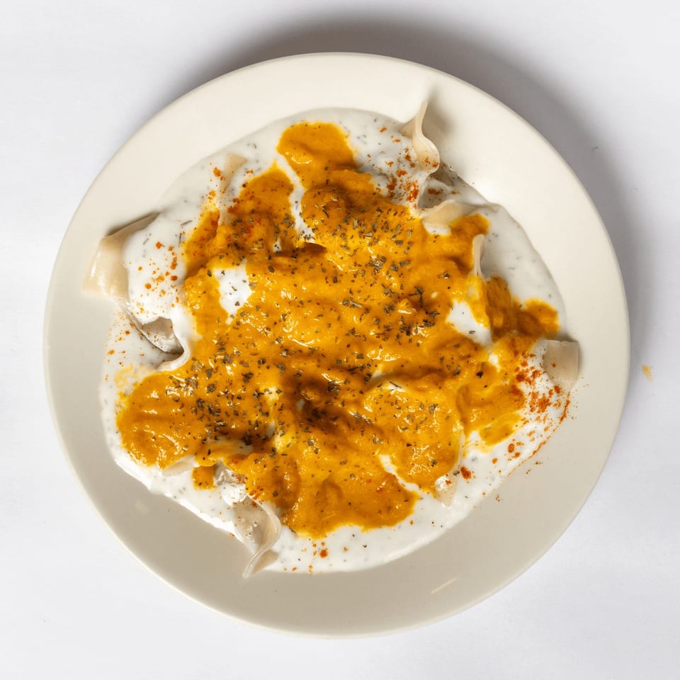 Overhead view of manti dumplings covered in white yogurt sauce and orange chili oil, garnished with herbs.