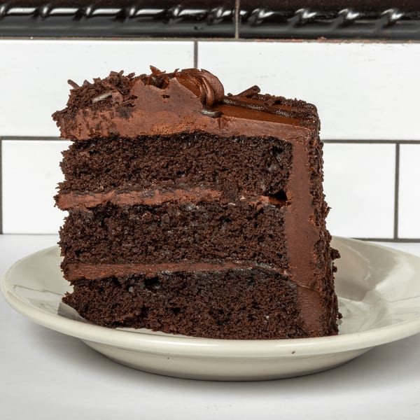 A rich slice of chocolate cake with chocolate frosting and chocolate shavings on a white plate.