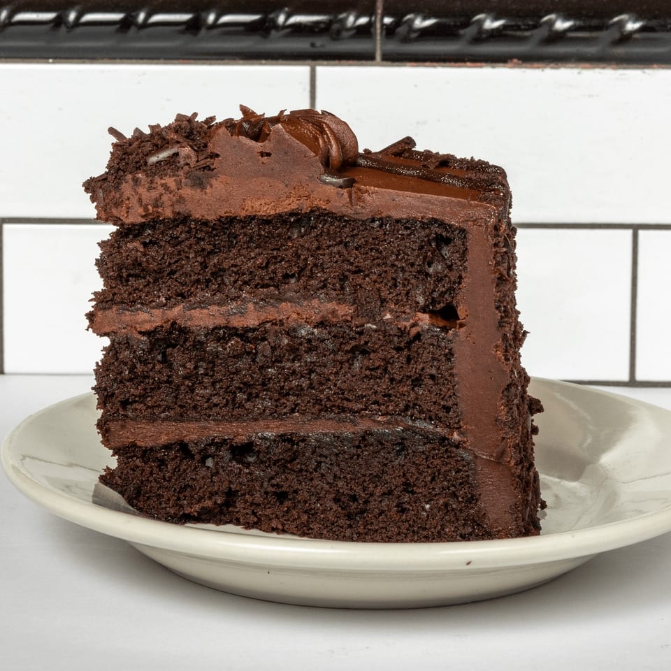 A rich slice of chocolate cake with chocolate frosting and chocolate shavings on a white plate.